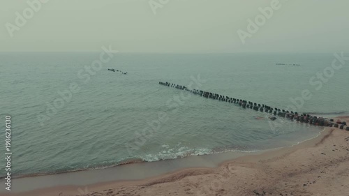 Wooden groynes and large rocks on the seashore. Coastal landscape with rough waves hitting barrier structures. Minimalist marine scenery for nature background.