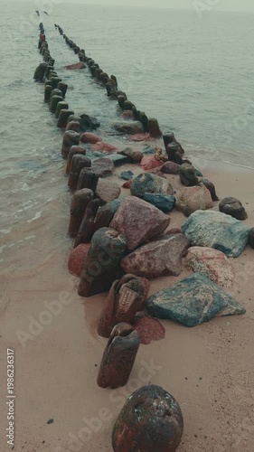 Wooden groynes and large rocks on the seashore. Coastal landscape with rough waves hitting barrier structures. Minimalist marine scenery for nature background.