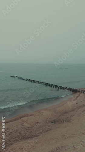 Wooden groynes and large rocks on the seashore. Coastal landscape with rough waves hitting barrier structures. Minimalist marine scenery for nature background.