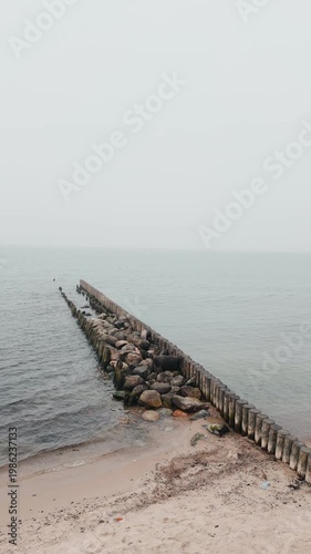 Wooden groynes and large rocks on the seashore. Coastal landscape with rough waves hitting barrier structures. Minimalist marine scenery for nature background.