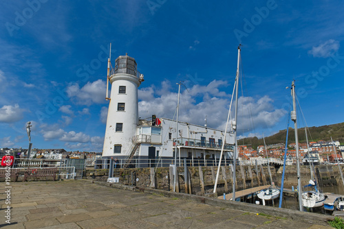 A tall white lighthouse and attached building overlook a bustling harbor with boats under a bright blue sky in Scarborough - Yorkshire - Great Britain