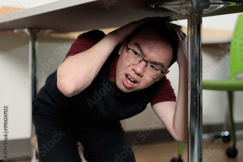 An Asian college student crouches under a desk with hands covering his head during an earthquake drill, for emergency preparedness, campus safety, or disaster training concepts.