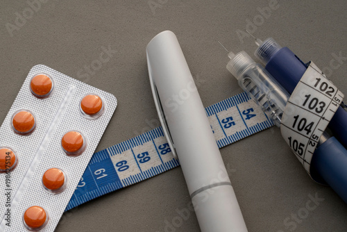 Medical weight loss concept with insulin pens, orange pills in blister pack, and measuring tape on a gray background for obesity treatment