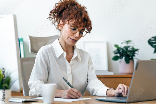 Concentrated mature business woman working with laptop while taking notes in the office