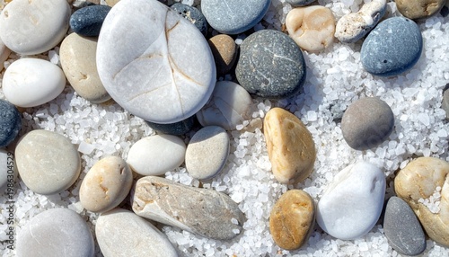 Naturally smooth and polished colorful pebbles of various sizes and shades resting on a coarse bright white sand beach creating a tranquil natural texture background