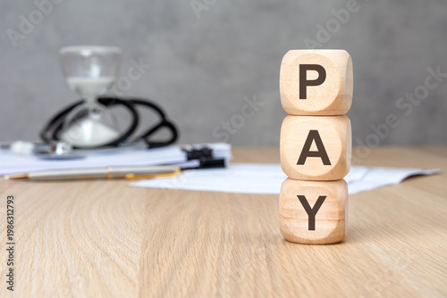 PAY word on vertically stacked wooden blocks. Blurred medical background with stethoscope and hourglass. Healthcare cost concept.