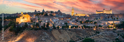 Panoramic shot captures the historic skyline of Toledo, Spain, at sunset, where the golden glow of the sky illuminates iconic landmarks like the Alcázar and the Primate Cathedral.