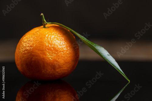 one fresh orange small tangerine with green leaves are reflected on a black mirror , a group of ripe sweet tangerines with green leaves with reflection on a black glass
