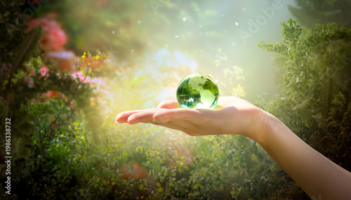 Earth Day or World Environment Day, environmental theme. Crystal glass globe ball in human hand on green fern fronds and tree leaves background. Save planet and protect nature, sustainability concept.