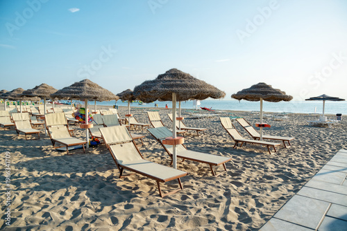 Lounge chairs with straw umbrellas on a sandy beach during the day by the ocean