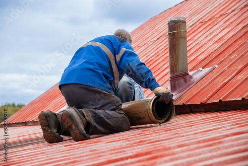 Chimney sweep worker installing pipe on red metal roof. Roofer fixing flue system on house exterior. Repair of heating ventilation and construction maintenance concept. Professional builder.