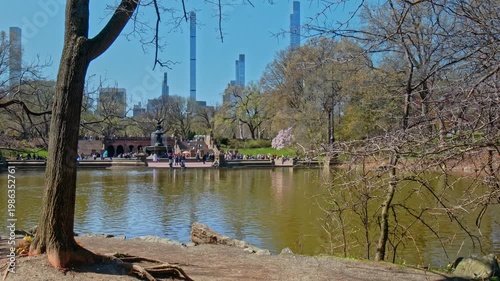 A 4K locked-off view of Bethesda Terrace and Fountain across the lake during early spring. Budding trees frame the iconic architecture and the massive pencil towers of the Midtown Manhattan skyline.