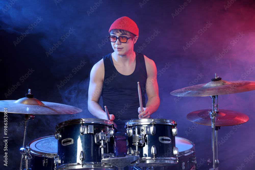 Obraz premium Teenage boy in glasses and hat learning how to play drums in color lights and smoke against dark background