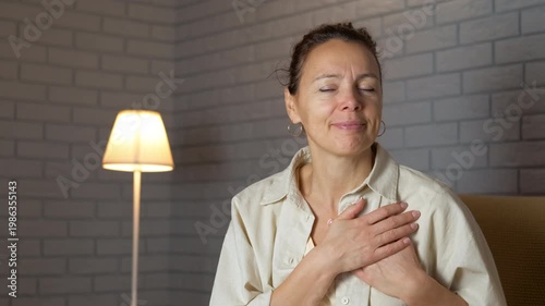 Grateful woman showing gratitude and love. Sincere woman sitting in a cozy room at night, putting her hands on her chest in a gesture of gratitude and appreciation while looking directly at the camera