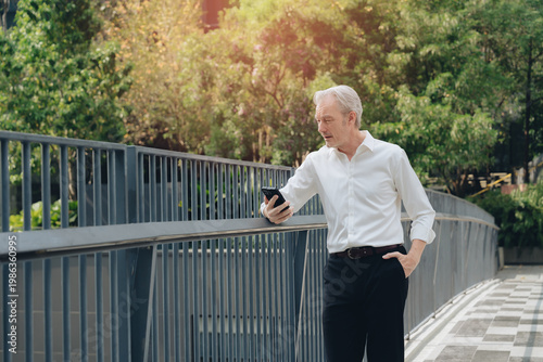 Middle aged man smartphone outdoor casual white shirt black pants railing trees sunlight urban setting