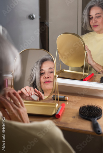 Senior woman doing her makeup, observing her reflection in a portable gold vanity mirror, focusing on self-care and beauty routine as she applies cosmetic products at a wooden dressing table