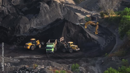 Heavy machinery including excavators and trucks is actively digging and transporting dark volcanic sand, creating large industrial piles within the Amed quarry in Bali. Aerial drone flight footage