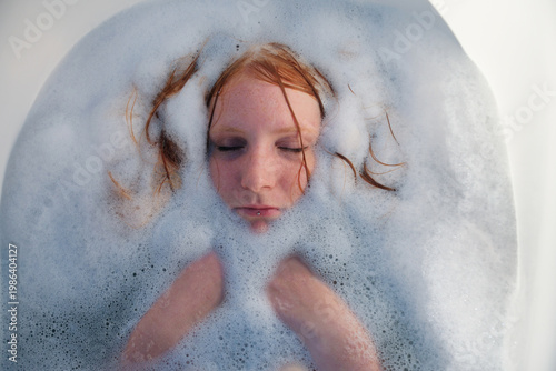 Woman relaxes in bath covered with foam with face emerging from bubbles eyes closed creating quiet intimate scene focused on cleansing rest and gentle sensory experience.