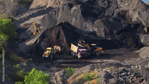 Construction vehicles wheel loaders and excavators moving and loading dark volcanic sand and rock into a dump truck, showcasing industrial mining operations in an open-pit quarry. Bali, Indonesia