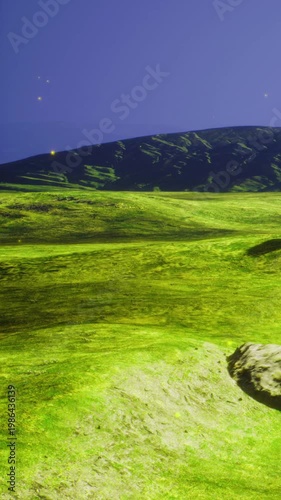 A large green field stretches under a clear sky. Mountains are visible in the background. The light from the setting sun creates a gentle glow. Small rocks are scattered throughout the field.