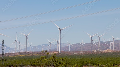 Multiple large white wind turbines spin in a field of windmills over a bright blue sky in the desert from ground level. 