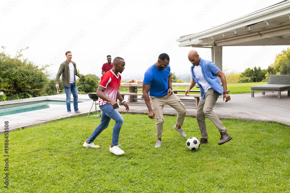Obraz premium Diverse male friends kicking soccer ball on backyard lawn beside picnic table and charcoal grill