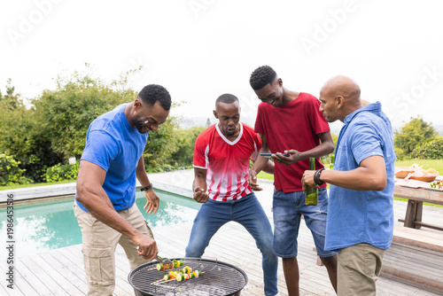 African American friends grilling kebabs on charcoal grill by pool, smartphone sparking cheering