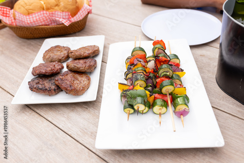 Vegetable skewers on wooden sticks resting on white platter at patio with burger patties