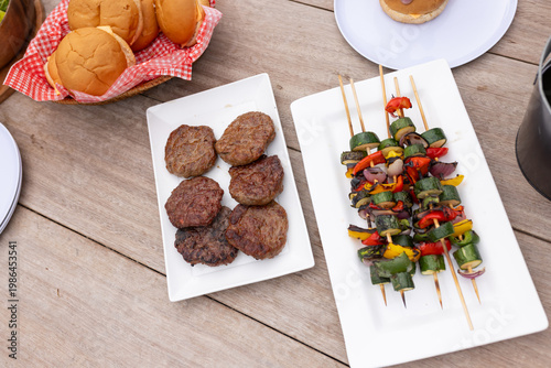 White plates sitting on picnic table holding six burger patties, vegetable skewers and bread basket