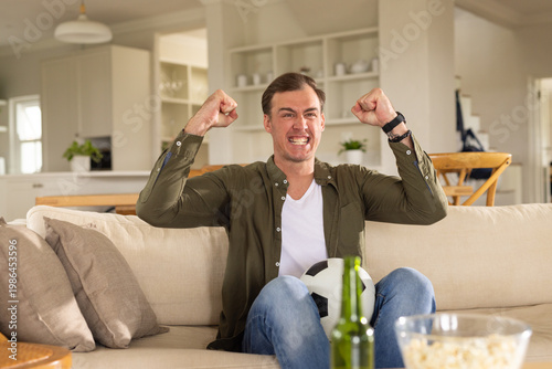 Adult man holding soccer ball on sofa with popcorn, reacting to TV goal, raising fists