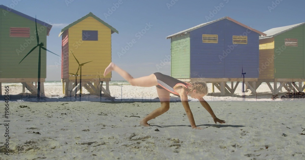 Obraz premium Cartwheeling girl wearing striped one-piece swimsuit on sandy beach, colorful huts on stilts