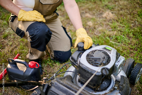 Unrecognizable gardener crouches to unscrew the fuel cap of a lawn mower tank, preparing to refill it with gasoline from a container during routine garden maintenance