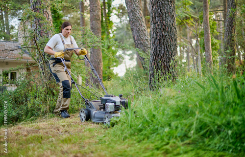 Young female gardener wearing work clothes and gloves pushes a gasoline lawn mower while cutting tall grass to tidy and maintain a large outdoor garden area.