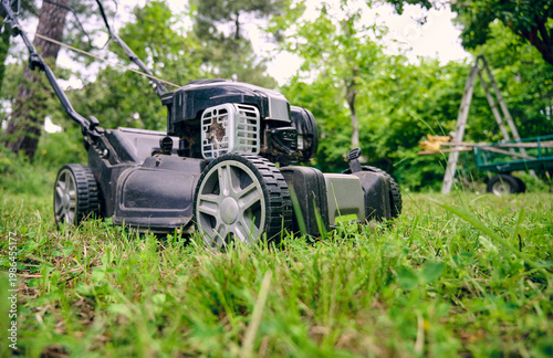 Close view of a powerful gasoline lawn mower resting on the grass of a rural garden before starting mowing work in an outdoor maintenance activity.