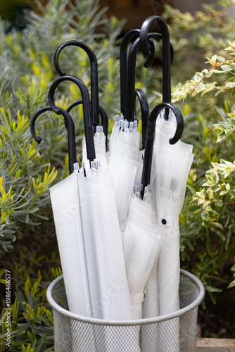 White umbrellas with black handles stored in a metal mesh stand, set against lush green garden shrubs. Clean, minimal, and elegant