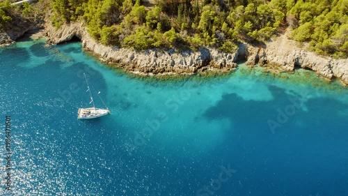 White sailboat anchored in a turquoise bay near a rocky forested coast in summer