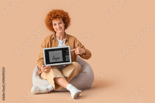Redhead female graphic designer pointing at laptop in beanbag on beige backgr...