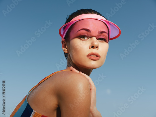 Close-up portrait of a young woman wearing a pink visor, sunlit skin and glossy lips, looking confidently toward the camera with a bold summer vibe and radiant glow.
