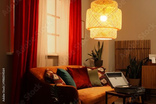 Comfortable sofa with modern laptop and glowing lamp near red curtains in living room
