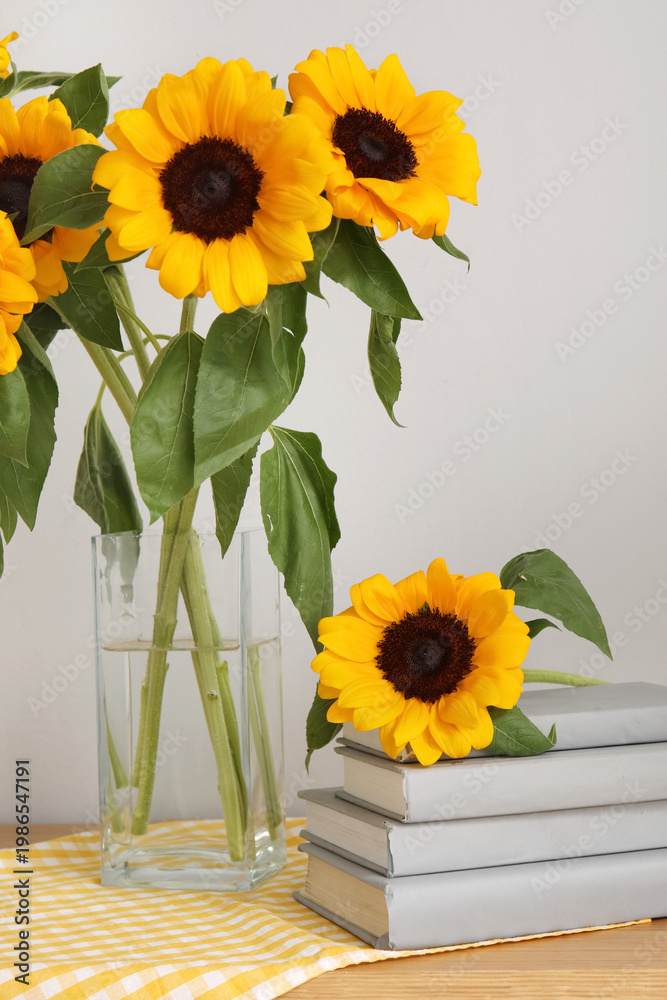 Fototapeta premium Vase with sunflowers and books on shelf near light wall