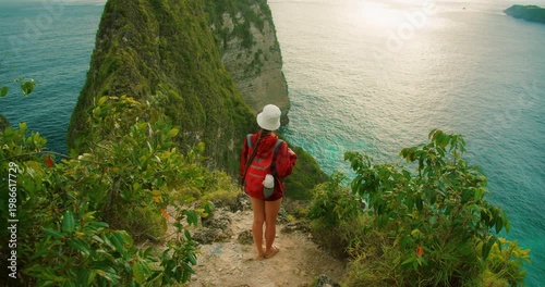 Woman with backpack looks at steep green cliff and blue ocean at Kelingking Nusa Penida Bali
