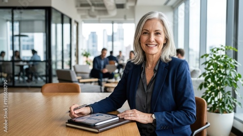 A mature woman sits at a wooden table in a modern office. She wears a navy blue blazer and a gray shirt.