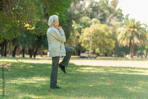 An elderly woman with gray hair practices a balance exercise, lifting one knee while standing on grass in a park setting