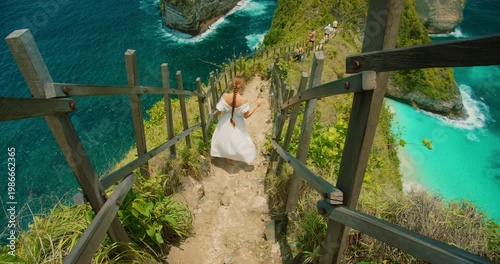 Woman in white dress walks down steep path at Kelingking Beach in Nusa Penida