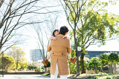 Rear View of a happy toddler girl being carried on her father's back in a public park, family bonding on a sunny winter day