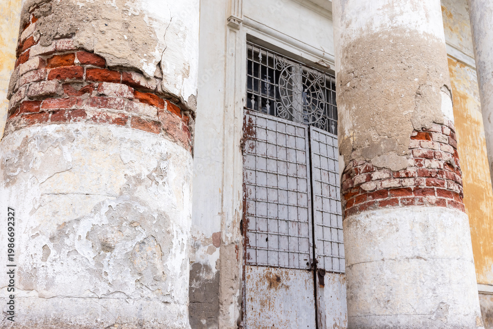 Fototapeta premium Weathered classical columns flank a rusted metal gate on a historic building, revealing exposed red brick, peeling plaster and textured decay that conveys age, neglect and architectural character