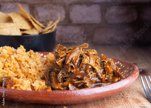 Pollo en mole, servido con arroz sobre un plato artesanal Mexicano en una mesa de madera con un tazon negro lleno de totopos o CHIPS de maiz en el fondo, con un muro rustico de ladrillos, 