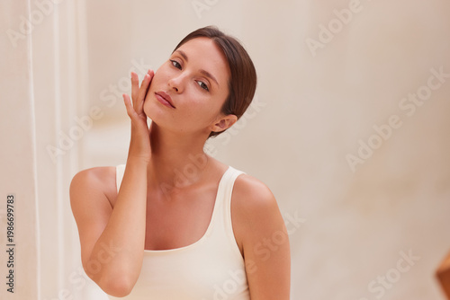 Natural beauty. Woman gently touching her cheek and jaw with one hand, looking sideways upward with a soft confident expression. Glowing bare skin, clean beige column background, copy space right.
