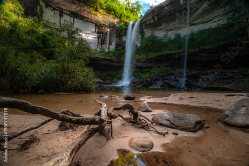 A waterfall cascading from a high cliff in the middle of the forest, with a sandy bottom and trees.