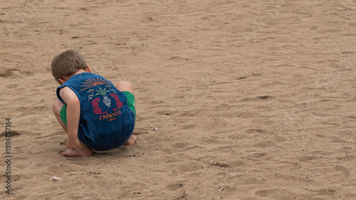 Photography a young boy seen from behind crouching and playing on a wide sandy beach during
